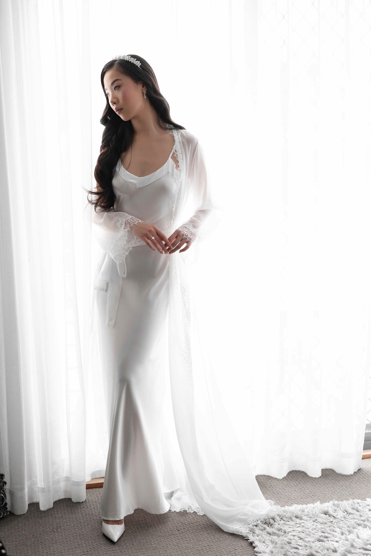 Bride wearing lace trim bridal robe with bridal tiara in front of white curtains