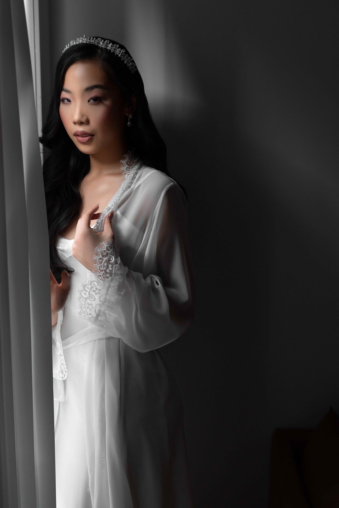Bride wearing lace trim bridal robe with bridal tiara next to window