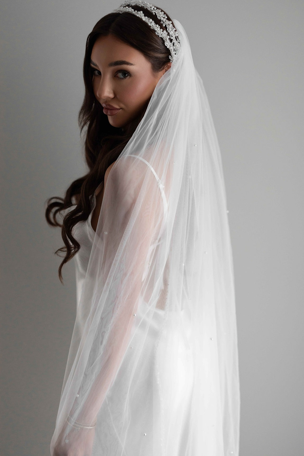Woman wearing a white wedding pearl veil and bridal headband against a gray background