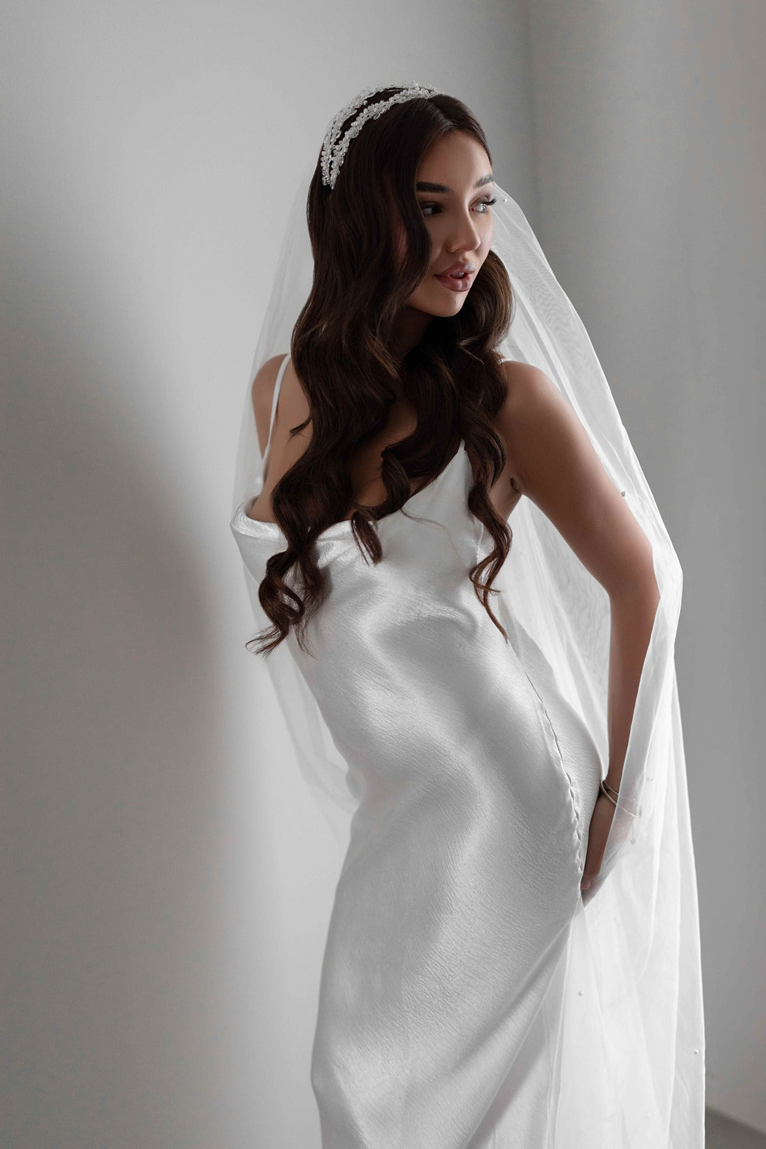 Bride in a white slipdress with a long pearl veil against a plain background