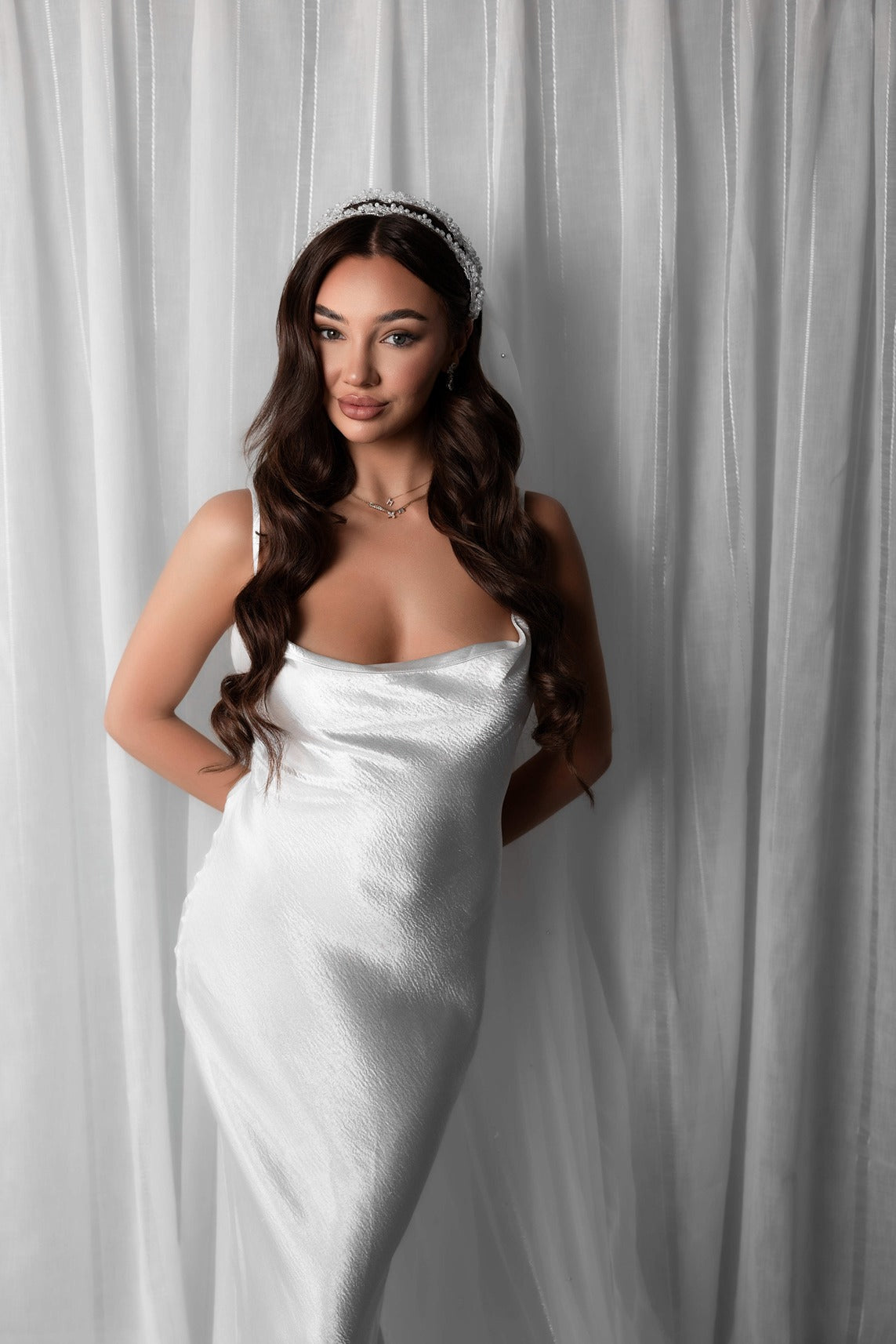 Woman in a white satin dress standing against a white curtain with a pearl veil