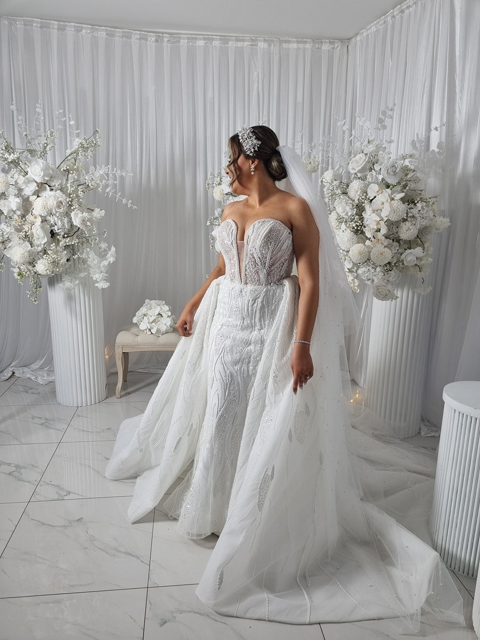 Woman in a white wedding dress and pearl veil standing in a decorated room with floral arrangements.