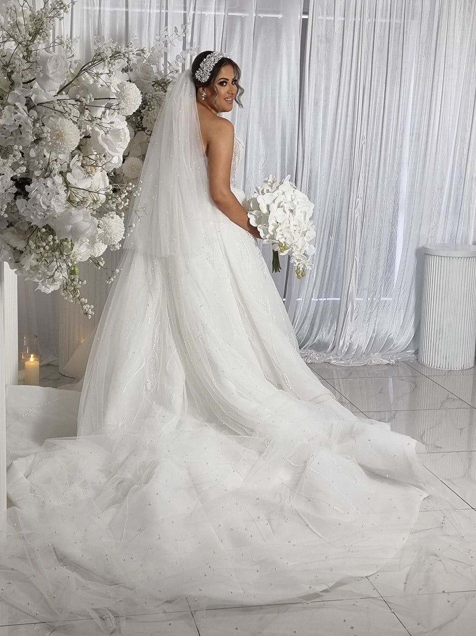 Bride in a pearl veil and white wedding dress holding a bouquet in a decorated room with white flowers and curtains.