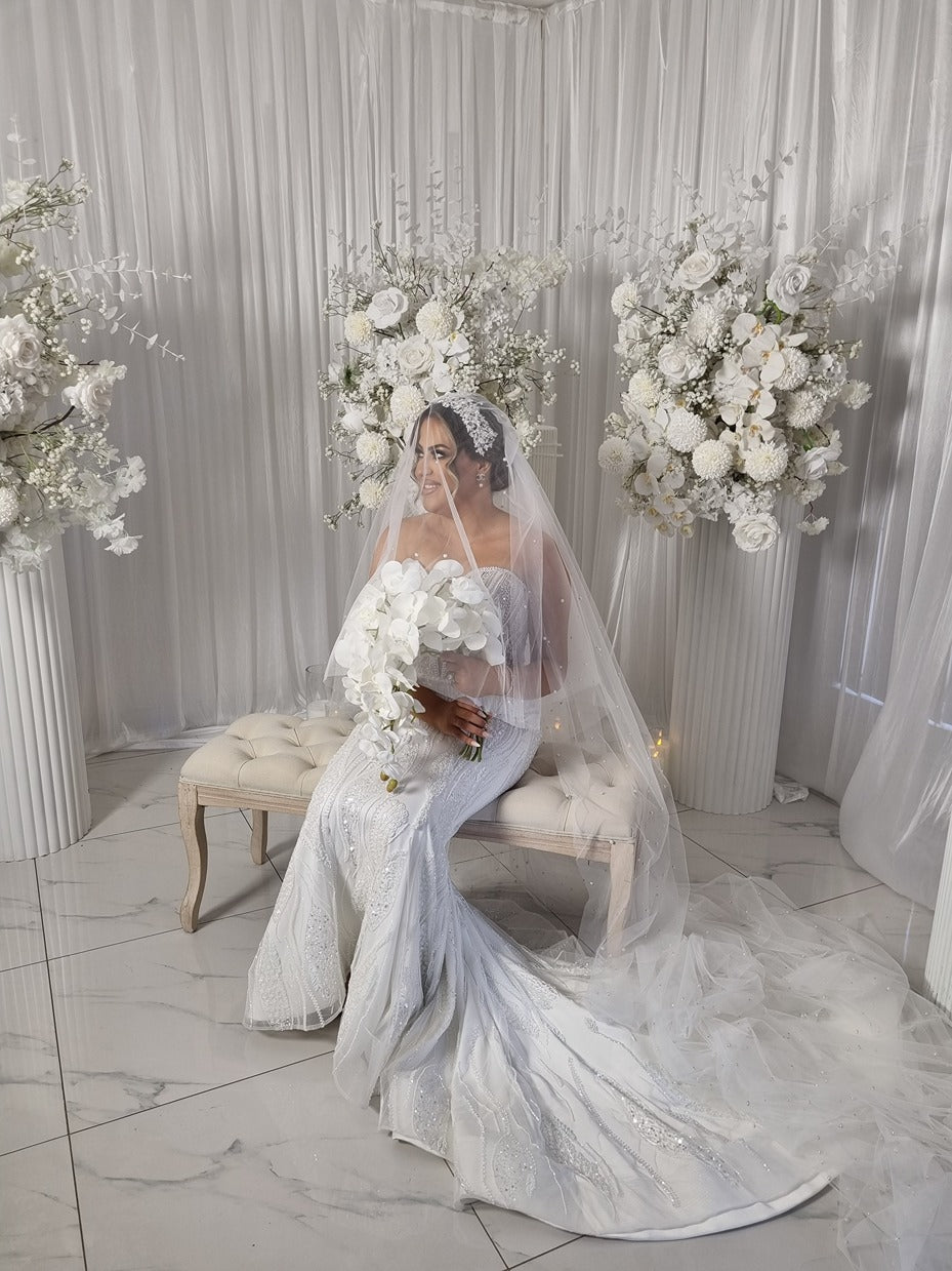 Woman in a white wedding dress and pearl veil standing in a decorated room with floral arrangements.