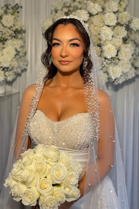 A bride wearing a strapless white beaded wedding gown with a pearl mantilla veil, holding a large bouquet of white roses against a backdrop of white floral arrangements