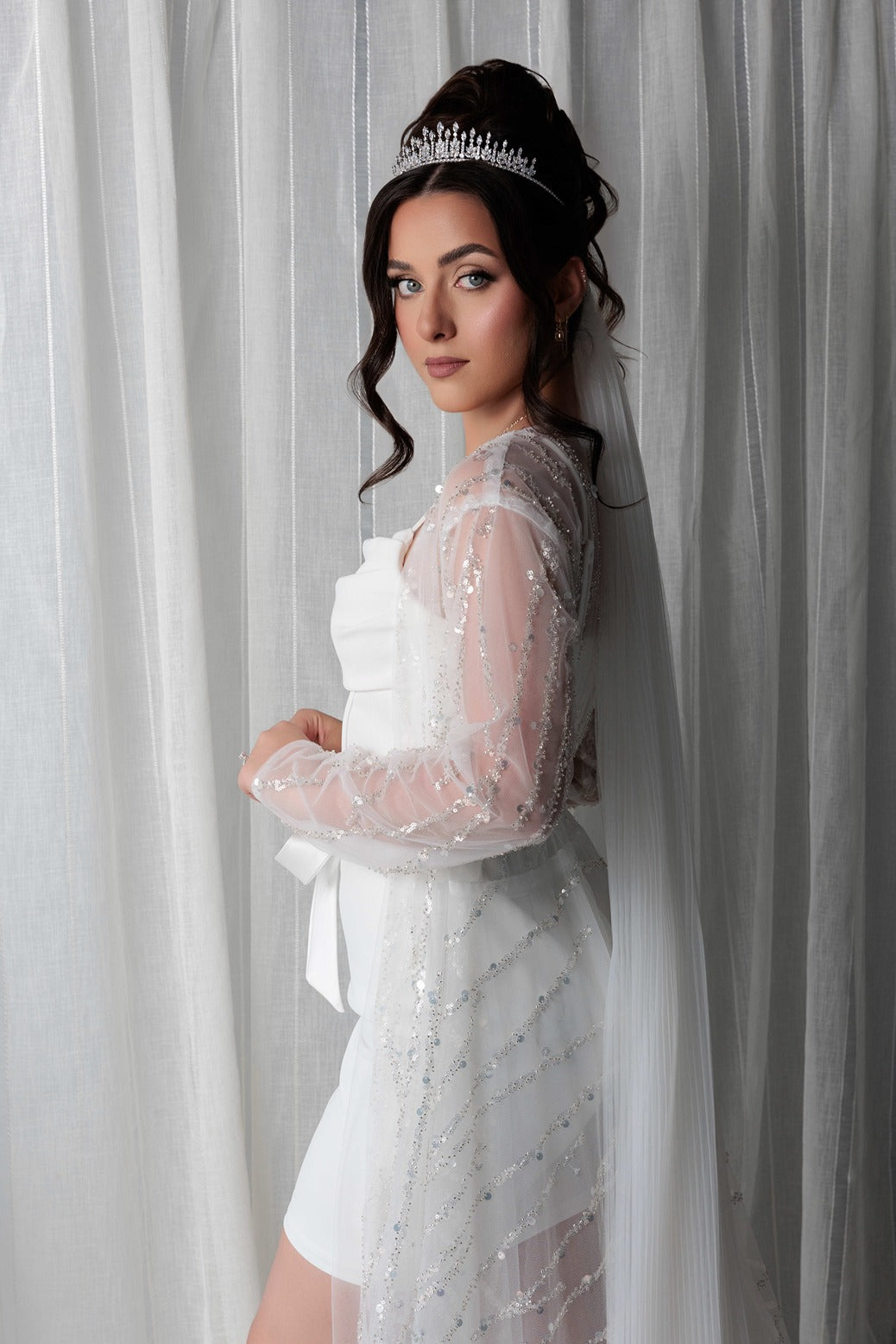 Woman in a bridal robe and a tiara, standing against a white curtain.