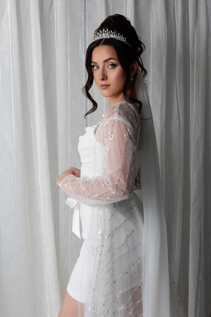 Woman in a bridal robe and a tiara, standing against a white curtain.