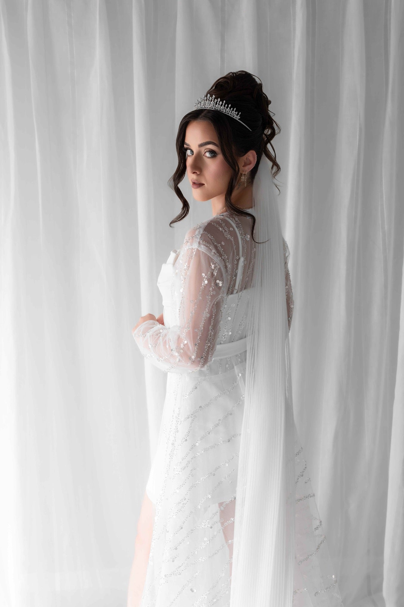 Woman in a luxury bridal robe and a tiara standing behind a white curtain.
