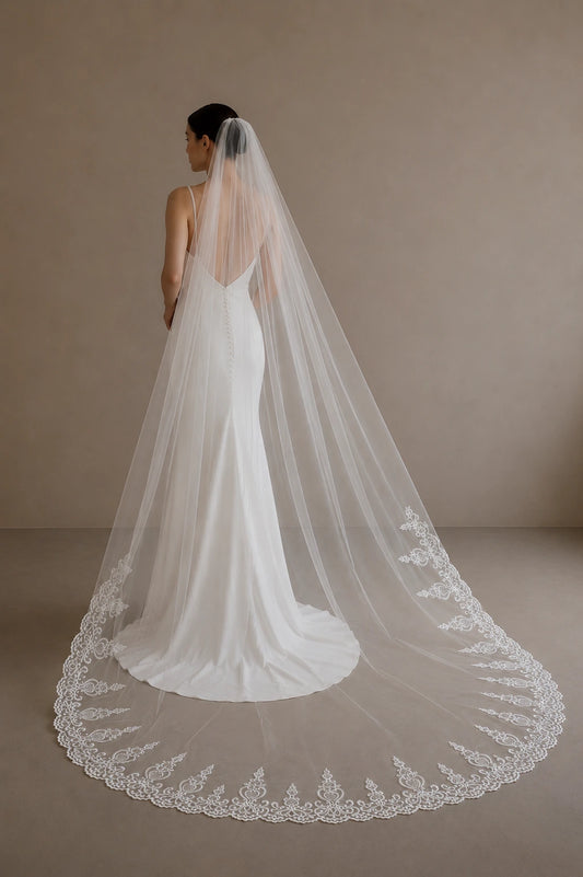 A woman wearing a white bridal gown and a very long sheer lace veil, photographed from the back against a plain warm beige studio backdrop.