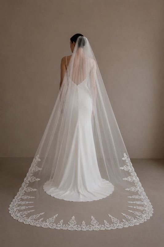 A woman seen from behind wearing a long white bridal veil with ornate lace trim over a white wedding dress, standing against a plain warm beige studio background.