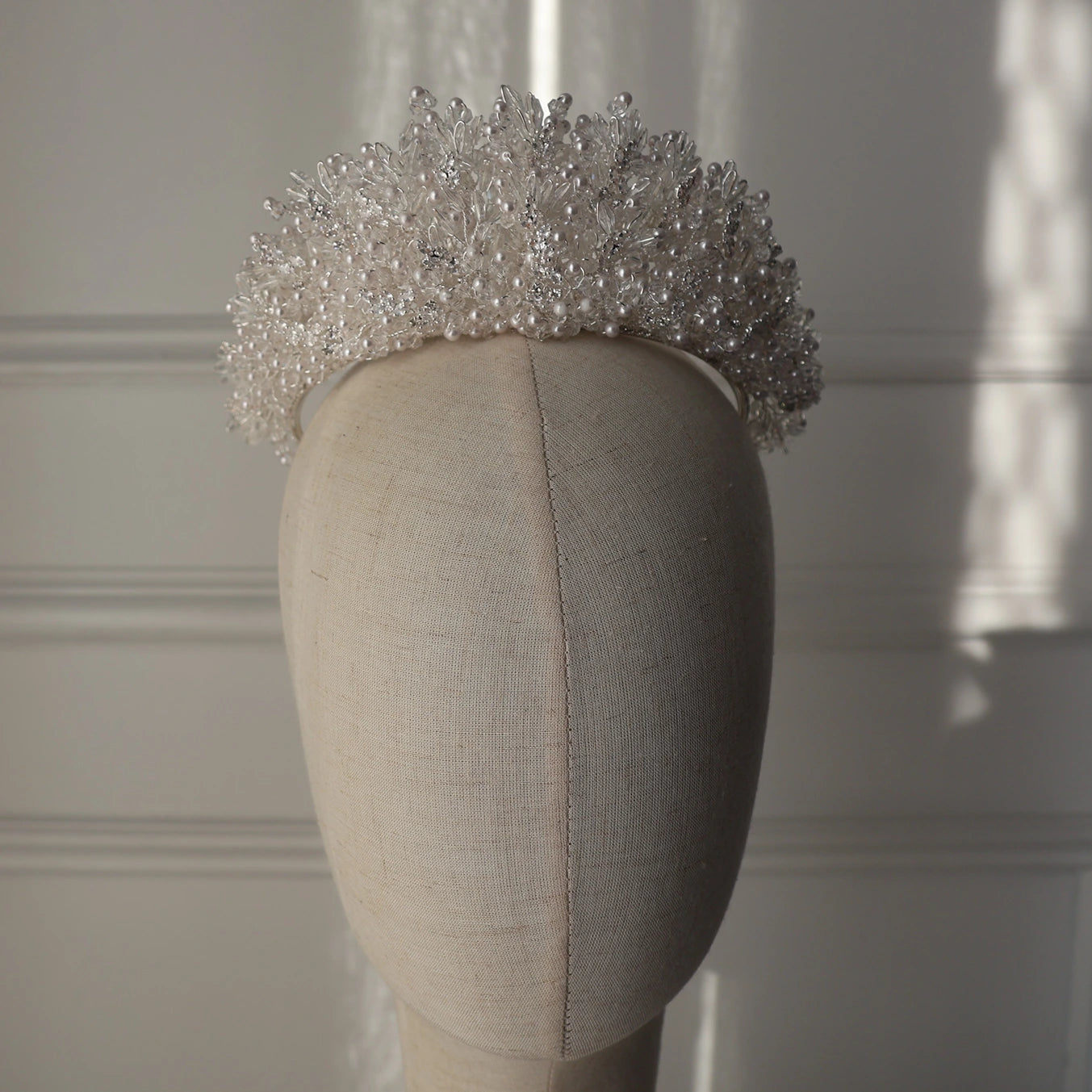 A silver beaded crown displayed on a beige fabric mannequin head against a white paneled wall with soft window light.