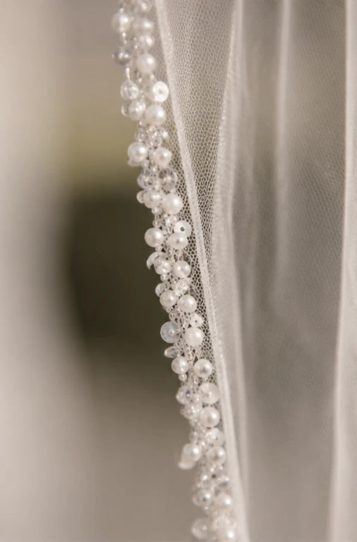 Close-up of a bridal veil with delicate pearl and crystal bead trim along the edge, shown against a softly blurred neutral background.