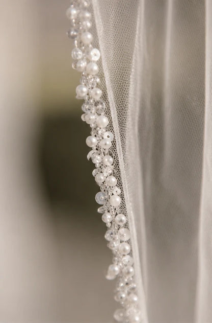 Close-up of a bridal veil with delicate pearl and crystal bead trim along the edge, shown against a softly blurred neutral background.