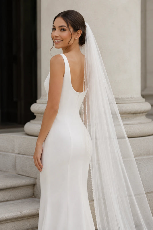 A woman wearing a fitted white wedding dress with a long pearl beaded edge 1 tier wedding veil, photographed from the back in front of a stone column and steps