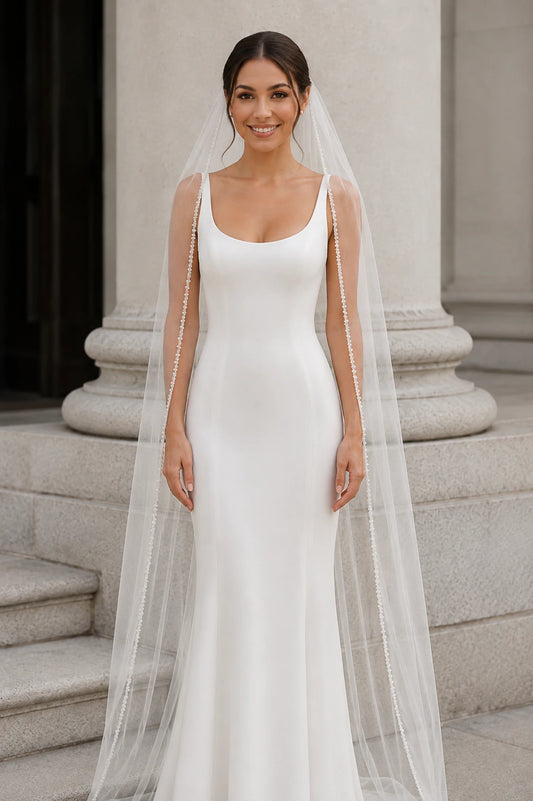 A woman wearing a fitted white wedding dress with a beaded pearl veil, standing on stone steps in front of a classical building.