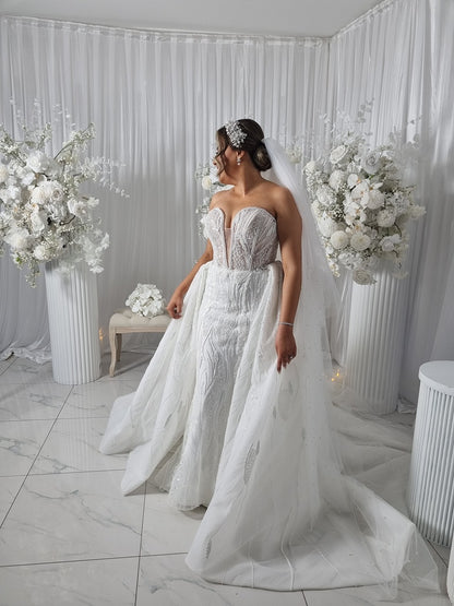 Woman in a white wedding dress and pearl veil standing in a decorated room with floral arrangements.