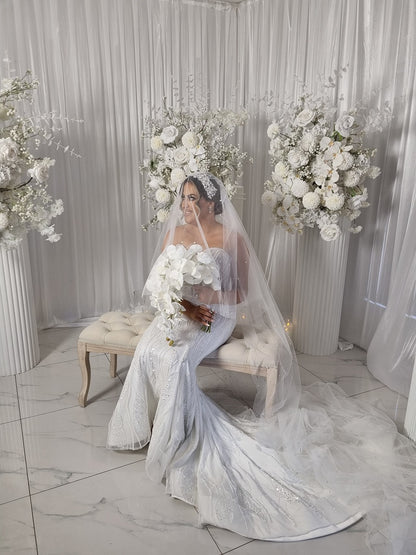 Woman in a white wedding dress and pearl veil standing in a decorated room with floral arrangements.