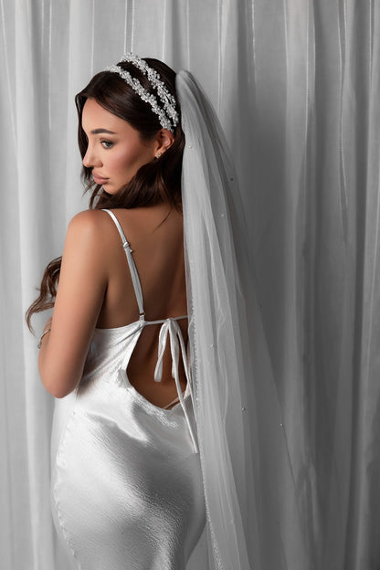Woman in a white bridal slip dress with a pearl veil and bridal headband against a white curtain background